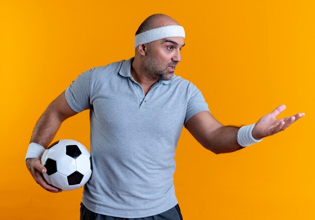 Hombre deportivo maduro en diadema sosteniendo un balón de fútbol mirando a un lado con el brazo extendido como pidiendo o discutiendo de pie sobre la pared naranja