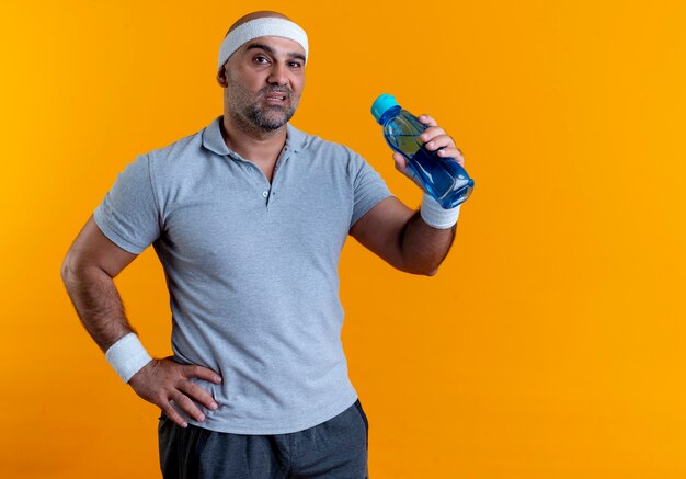 Hombre deportivo maduro en diadema sosteniendo una botella de agua mirando hacia el frente con cara seria de pie sobre la pared naranja