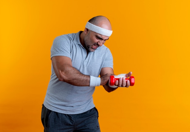 Hombre deportivo maduro en diadema tocando su mano vendada buscando malestar que sufren de dolor de pie sobre la pared naranja
