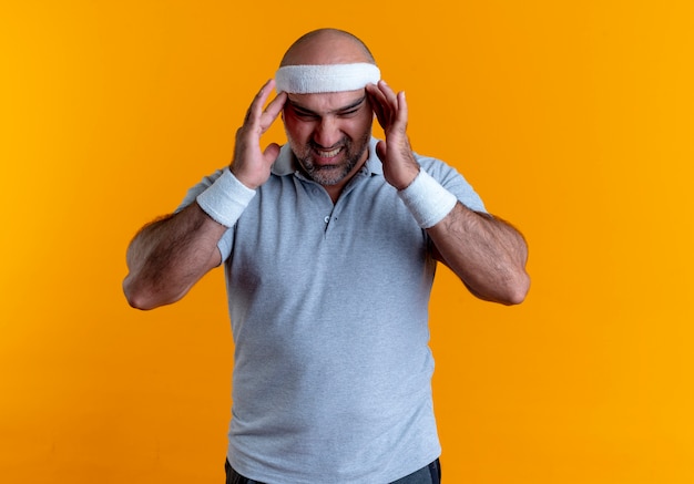 Hombre deportivo maduro en diadema tocando templos que sufren de fuertes dolores de cabeza de pie sobre la pared naranja