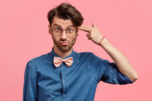 Hombre deprimido, sin afeitar y disgustado con un corte de pelo moderno, finge suicidarse, dispara con el dedo índice en la sien, vestido con camisa vaquera y corbatín rosa, se siente cansado de la vida difícil. Negatividad