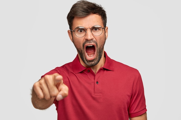 Hombre deprimido furioso con cerdas oscuras, le grita enojado a alguien, puntos, vestido con camiseta roja brillante, aislado sobre una pared blanca. Un loco sin afeitar expresa rabia, grita fuerte