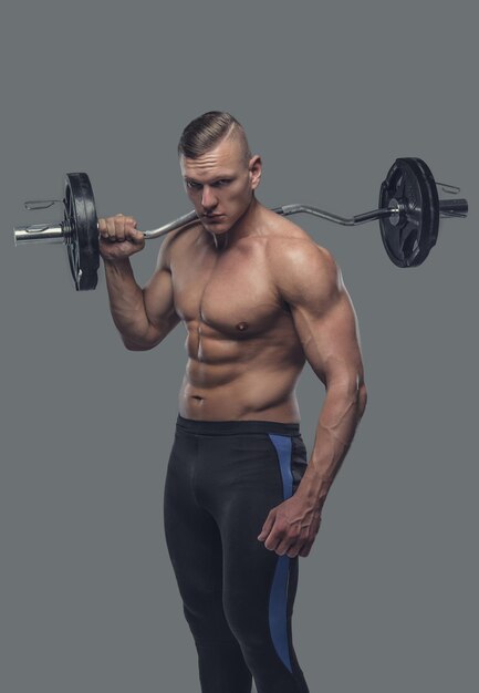 Hombre descamisado muscular que sostiene la barra en su hombro. Aislado en un fondo gris.