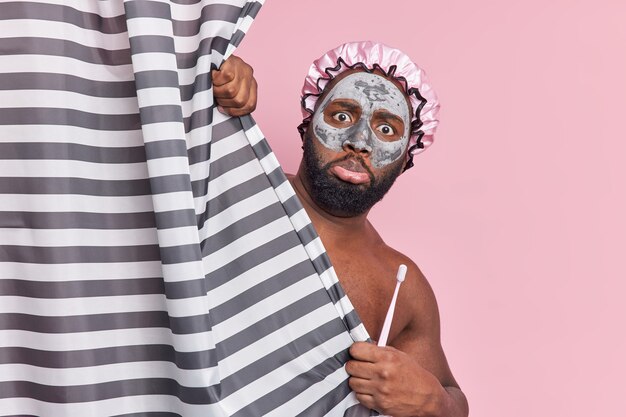 Hombre desconcertado con barba espesa aplica máscara de arcilla nutritiva usa gorro de baño sostiene cepillo de dientes esconde cuerpo desnudo detrás de cortina de ducha se somete a procedimientos de higiene diaria aislados en pared rosa