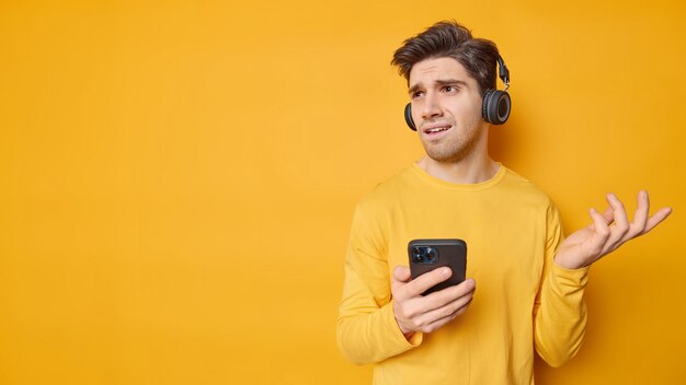 El hombre desconcertado y disgustado se encoge de hombros y mira con insatisfacción lejos, no puede descargar la canción a la lista de reproducción, usa auriculares en los oídos para escuchar poses de música contra el espacio de copia de fondo amarillo