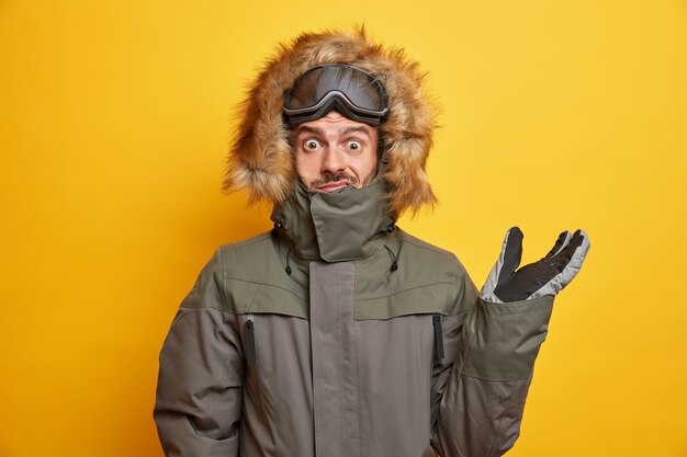 Hombre desconcertado en traje de invierno levanta la mano y parece confundido usa capucha de su abrigo anorak va a esquiar durante el tiempo libre.