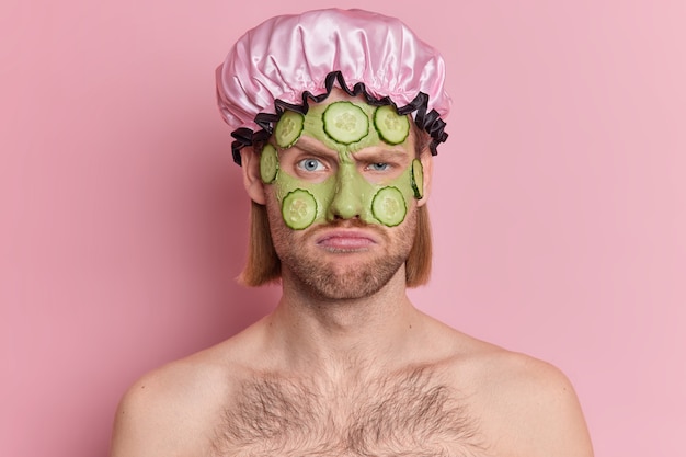 El hombre descontento frunce el ceño y mira infelizmente aplica máscara facial verde con rodajas de pepino quiere tener una piel sana usa sombrero de baño sin camisa.
