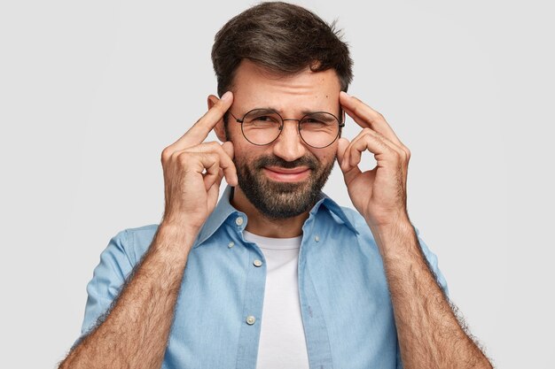 Hombre desesperado sin afeitar mantiene las manos en las sienes, frunce el ceño con disgusto, sufre de dolor de cabeza, vestido informalmente aislado sobre una pared blanca. Hombre guapo expresa frustración, sentimiento negativo