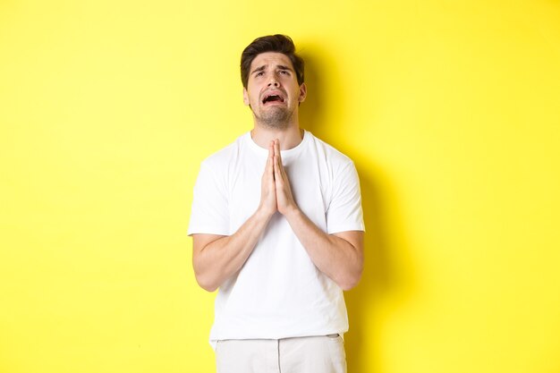 Hombre desesperado suplicando a Dios, tomados de la mano en oración y mirando hacia arriba angustiado, de pie sobre un fondo amarillo. Copia espacio