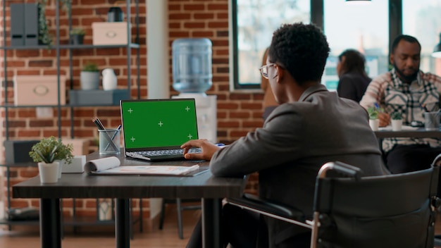 Hombre con discapacidad mirando la pantalla verde en la computadora, sentado en silla de ruedas. Trabajador de oficina que usa una plantilla aislada con cromakey en blanco y fondo de espacio de copia en la pantalla del portátil.