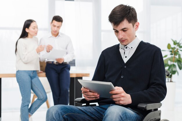 Hombre discapacitado joven que usa la tableta digital a disposición que se sienta en la silla de ruedas con su colega en el fondo