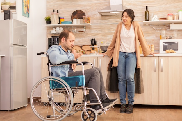 Hombre discapacitado en silla de ruedas mirando ensalada hecha por esposa en la cocina. Hombre discapacitado paralítico discapacitado con discapacidad para caminar que se integra después de un accidente.