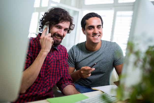 Hombre diseñador gráfico hablando por teléfono móvil mientras compañero de trabajo que mira el ordenador