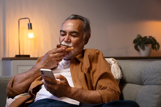 Hombre disfrutando de algo de comida mientras está solo en casa y usa un teléfono inteligente