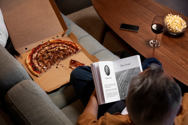 Hombre disfrutando de su tiempo a solas en casa mientras lee un libro y come pizza