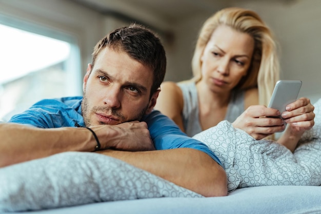 Hombre disgustado acostado en la cama e ignorando a su novia que está enviando mensajes de texto por teléfono móvil