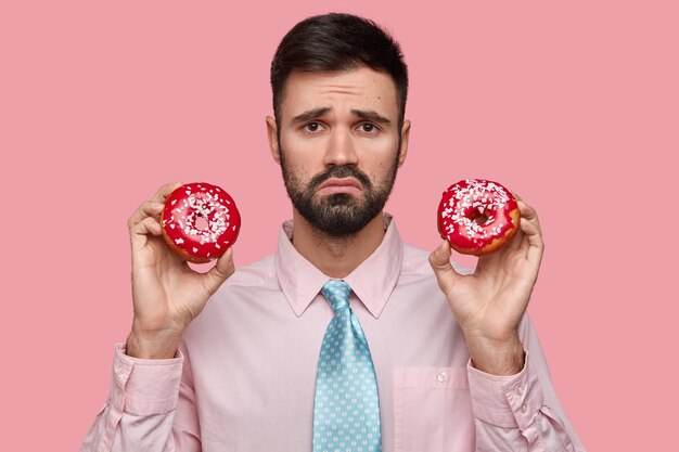 Hombre disgustado con barba oscura, ceño fruncido, sostiene dos deliciosas rosquillas, se siente infeliz porque no puede comer dulces