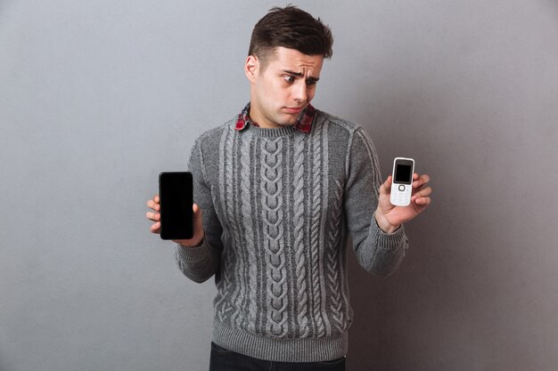 Hombre disgustado en suéter eligiendo entre teléfonos inteligentes y mirando