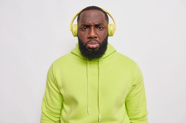 El hombre disgustado tiene una expresión de cara enfurruñada escucha la pista de audio a través de auriculares inalámbricos viste una cómoda sudadera verde aislada sobre una pared blanca