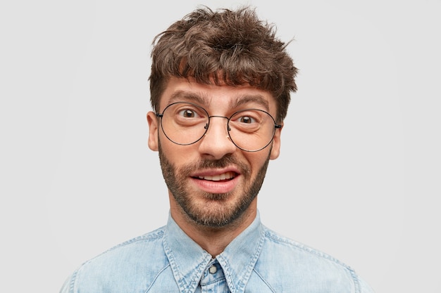 Hombre divertido con barba incipiente, tiene mirada indecisa y curiosa, frunce el ceño, mira directamente a la cámara, vestido con camisa de mezclilla, aislado sobre una pared blanca