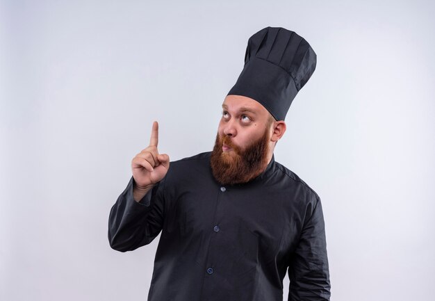 Un hombre divertido chef barbudo en uniforme negro apuntando hacia arriba con el dedo índice sobre una pared blanca