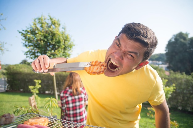 Hombre divertido en la fiesta familiar de BBQ. Hombre de cabello oscuro con camiseta amarilla comiendo carne recién asada y haciendo cara graciosa. Barbacoa, cocina, comida, concepto familiar.