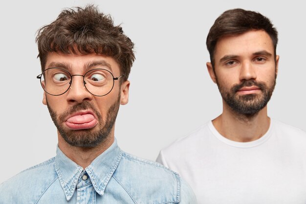 El hombre divertido hace muecas, hace tonterías, cruza los ojos y muestra la lengua, su amigo barbudo está en la pared, tiene una expresión seria