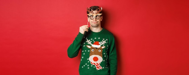 Foto gratuita hombre divertido en suéter de navidad y máscara de fiesta celebrando las vacaciones de invierno mostrando caras graciosas standi...