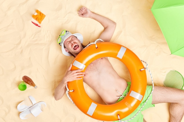 El hombre se divierte se encuentra en la arena blanca con un aro salvavidas naranja inflado en el cuerpo viste un sombrero blanco, máscara de snorkel, grita en voz alta, pasa el tiempo libre en la playa durante el caluroso día de verano