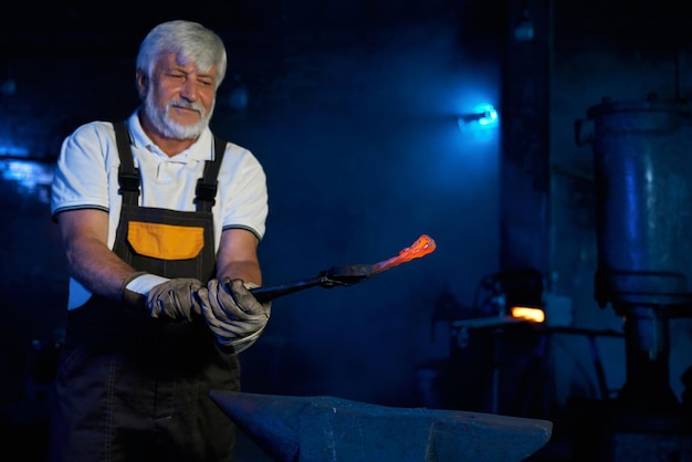 Hombre de edad caucásica en delantal de seguridad y guantes sosteniendo fórceps industriales con acero calentado Herrero competente preparando metal para procesar en yunque