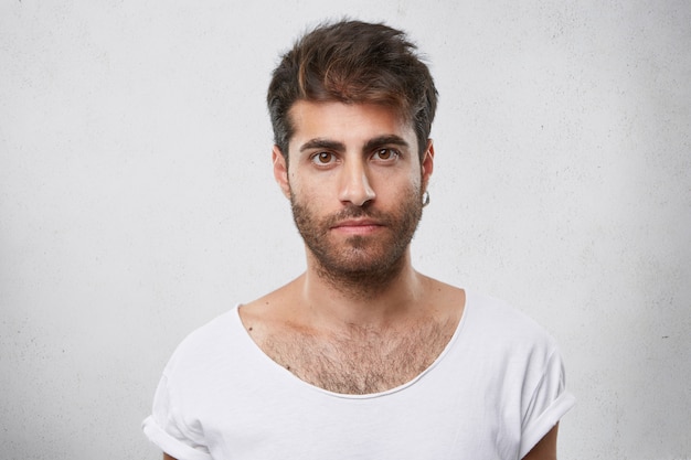 Hombre elegante con barba, peinado de moda, pendiente en la oreja y camiseta blanca mirando directamente con sus ojos oscuros.