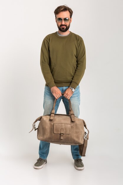 Hombre elegante barbudo confiado hermoso en sudadera con bolsa de viaje, jeans y gafas de sol aislados