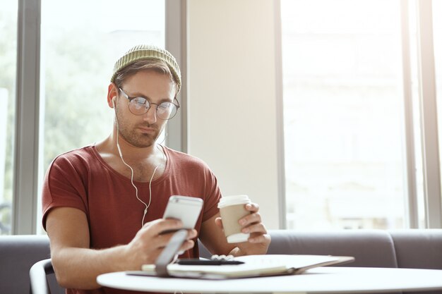 Hombre elegante barbudo viste camiseta roja casual y gafas, sostiene un teléfono celular genérico