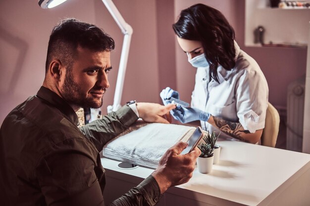 Un hombre elegante con camisa sosteniendo un teléfono y mirando de lado mientras recibe una manicura de una esteticista en el salón de belleza