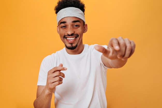 Hombre elegante con camiseta blanca se ríe y señala con el dedo a la cámara Retrato de un chico moreno con una diadema blanca sonriendo y bailando sobre fondo naranja