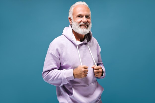 Hombre elegante con capucha blanca sonriendo Chico adulto de pelo gris de moda con barba con tatuajes en sudadera lilas mirando a la cámara