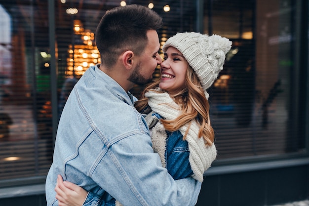 Hombre elegante en chaqueta de mezclilla abrazando a su novia en la calle urbana. Feliz pareja caucásica posando en la calle durante la fecha.