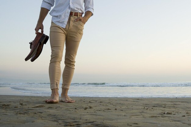 Hombre elegante con clase en camisa blanca y pantalones beige caminando en la playa con sus zapatos en las manos