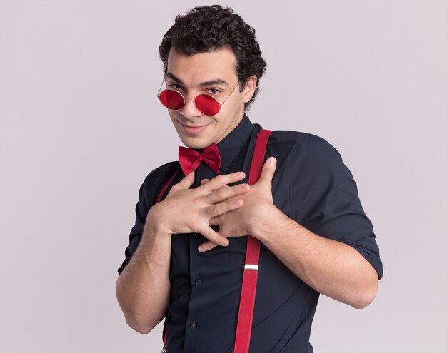 Hombre elegante complacido con pajarita con gafas y tirantes mirando al frente con las manos cruzadas sobre el pecho sintiéndose agradecido de pie sobre la pared blanca