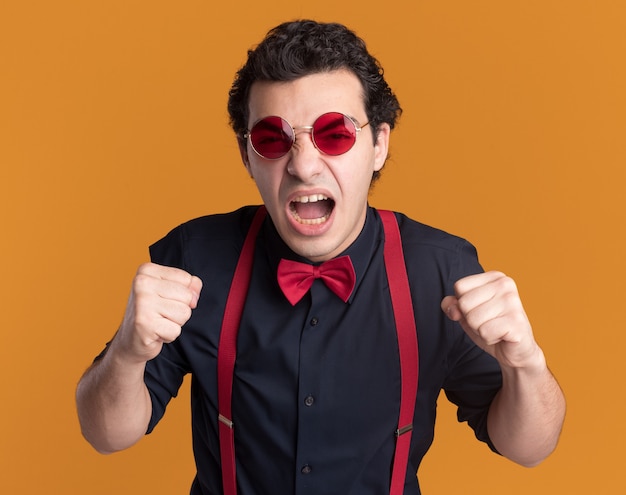 Hombre elegante enojado con pajarita con gafas y tirantes gritando puños apretados loco loco parado sobre pared naranja