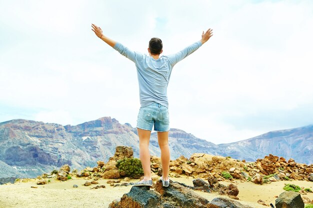 Hombre elegante feliz en ropa casual hipster de pie en el acantilado de la montaña con las manos levantadas al sol y celebrando el éxito