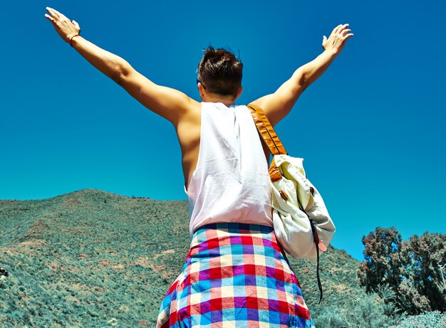 Hombre elegante feliz en ropa casual hipster saltando frente a la montaña con las manos levantadas al sol y celebrando el éxito