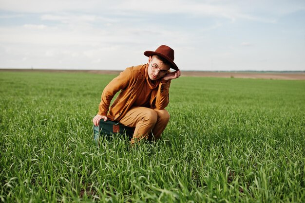 Hombre elegante con gafas, chaqueta marrón y sombrero con bolso posado en campo verde