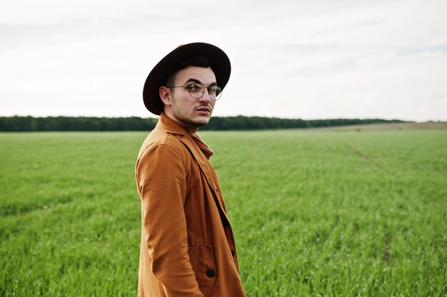 Hombre elegante con gafas, chaqueta marrón y sombrero posado en campo verde