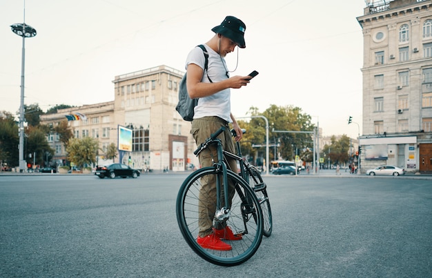 Hombre elegante joven que manda un SMS en el teléfono mientras que se coloca a un lado en bicicleta.
