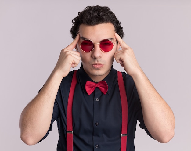 Hombre elegante con pajarita con gafas y tirantes con aspecto cansado y aburrido tocando sus sienes de pie sobre la pared blanca