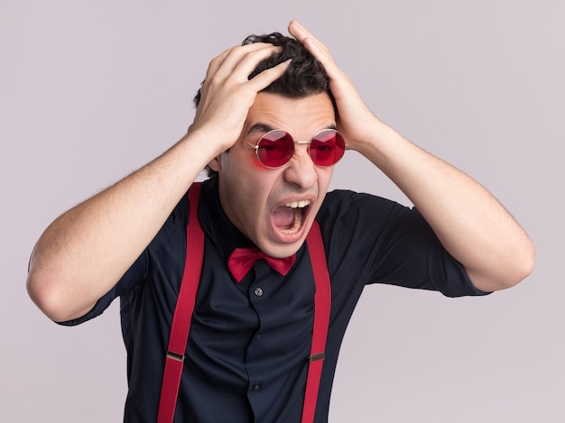 Hombre elegante con pajarita con gafas y tirantes gritando con expresión agresiva volviéndose loco con las manos en la cabeza de pie sobre la pared blanca