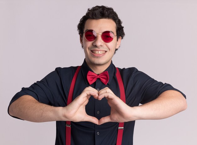 Hombre elegante con pajarita con gafas y tirantes haciendo gesto de corazón con los dedos sonriendo alegremente de pie sobre la pared blanca