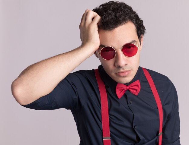 Hombre elegante con pajarita con gafas y tirantes mirando al frente confundido con la mano en la cabeza por error de pie sobre la pared blanca