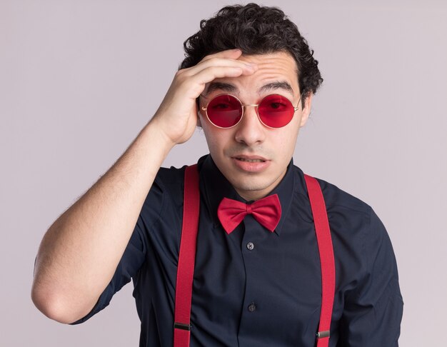 Hombre elegante con pajarita con gafas y tirantes mirando al frente confundido con la mano en la frente de pie sobre la pared blanca